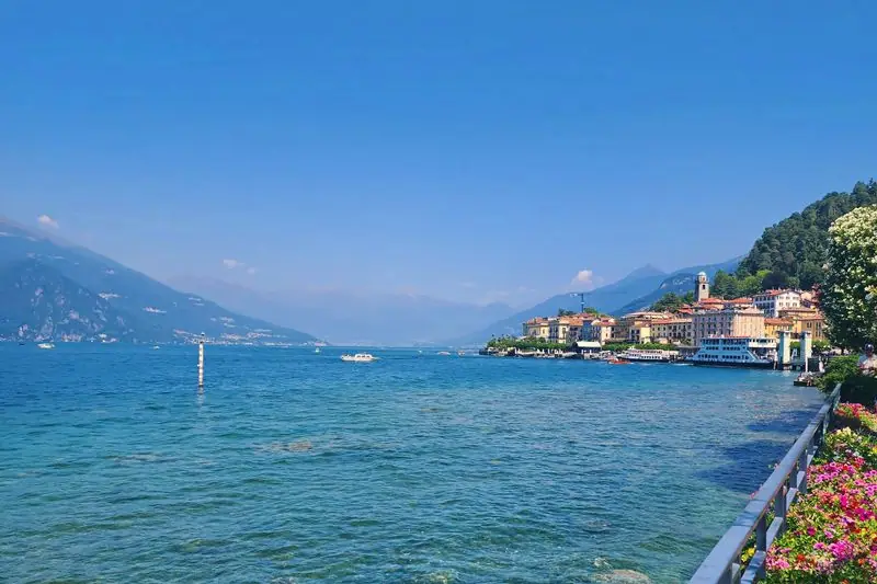 The Bellagio waterfront on Lake Como, pastel houses and a ferry quay above clear green shallows, mountains beyond