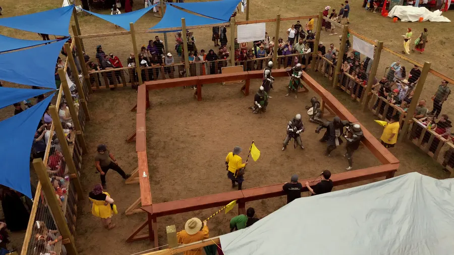 Aerial view of the timber combat pen under blue shade sails — armored team bouts, spectators packed at the fence