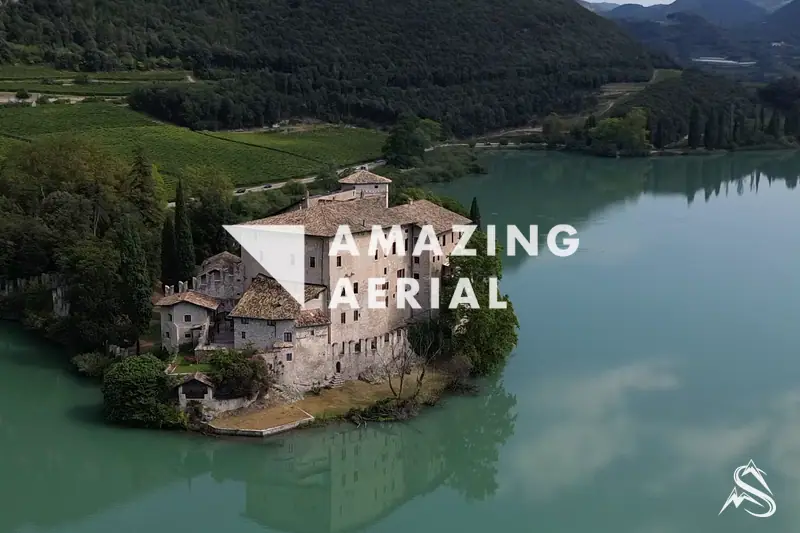 Aerial of Castel Toblino on its wooded spit, vineyards and cypresses behind it above the green lake