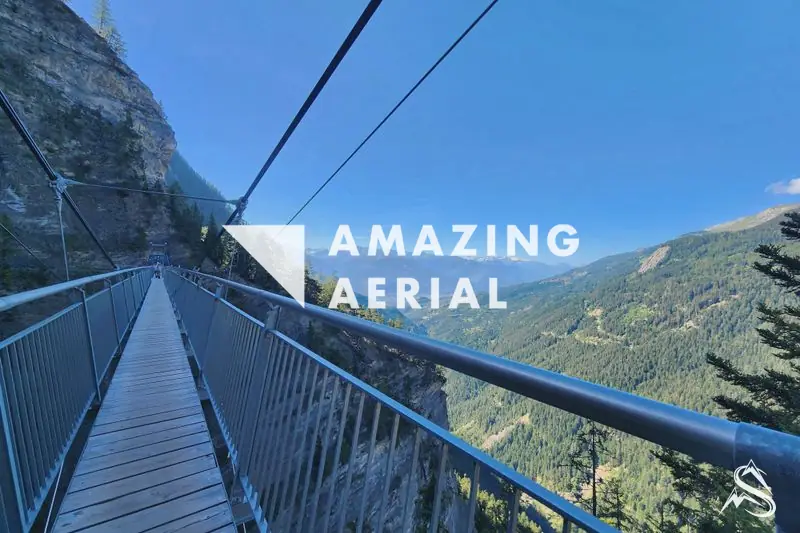 A suspension footbridge running out across a wooded gorge above the Rhône valley, cables rising to the far side