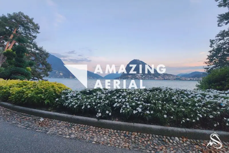 The Lugano lakefront promenade at dusk, white flowerbeds and a church tower framed by a tree, Monte San Salvatore across the water