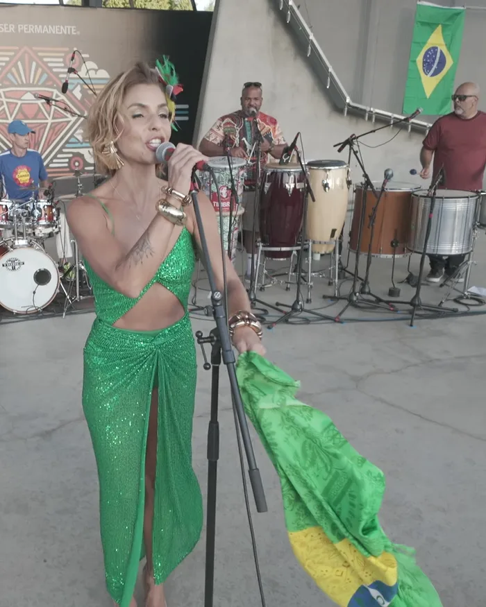 A singer in a green dress performing in front of the full percussion band, a Brazilian flag and the venue's sponsor signage behind her