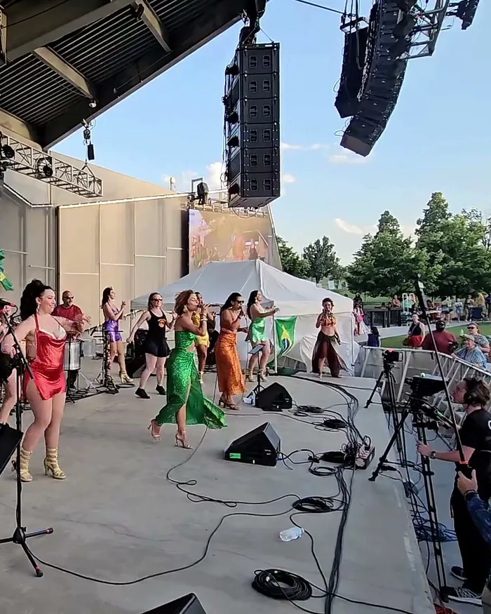 A line of dancers and singers in red, purple, green, and orange performance outfits clapping and dancing together in front of the band