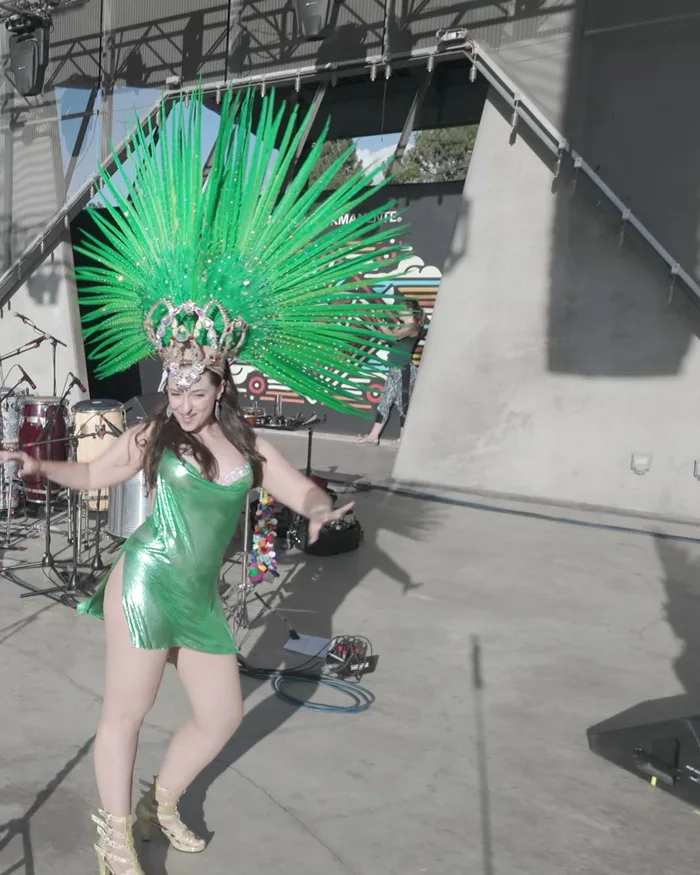 A dancer in a green feathered headdress mid-dance, smiling, fully in frame on the stage apron