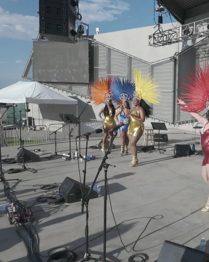 Four dancers in orange, blue, yellow, and red feathered headdresses on stage, the center dancer in blue smiling directly at the camera