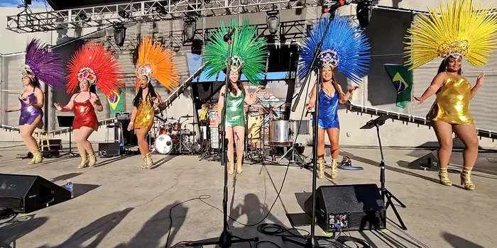 All six Boulder Samba School dancers lined up on stage in purple, red, orange, green, blue, and gold feathered headdresses, a wide establishing view of the full line