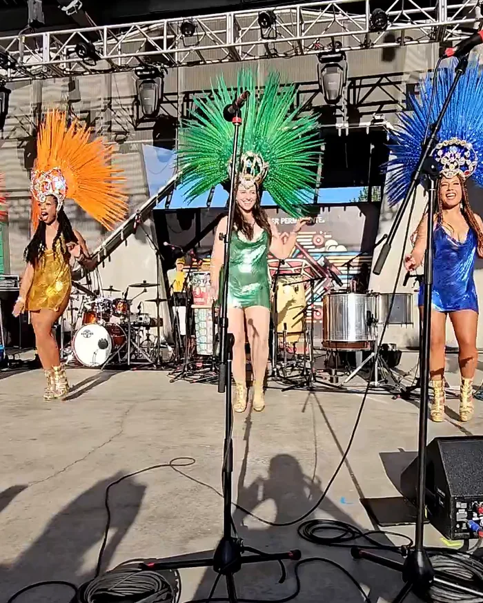 Three Boulder Samba School dancers in orange, green, and blue feathered headdresses, all smiling and facing the camera mid-song, the percussion band behind them