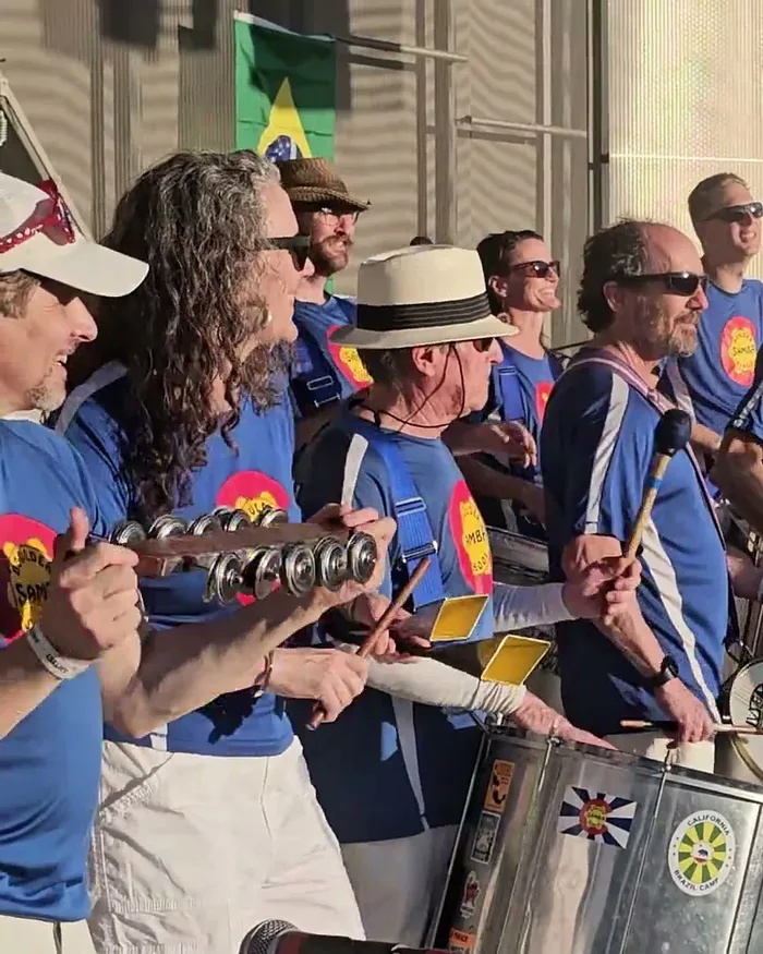 Boulder Samba School percussionists in matching blue shirts playing congas and surdos, several faces visible and smiling