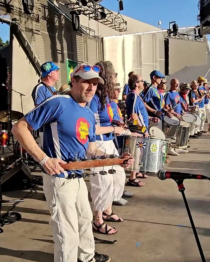 Boulder Samba School's full percussion line on stage, their shirts reading the group's name over a Colorado-flag-style patch