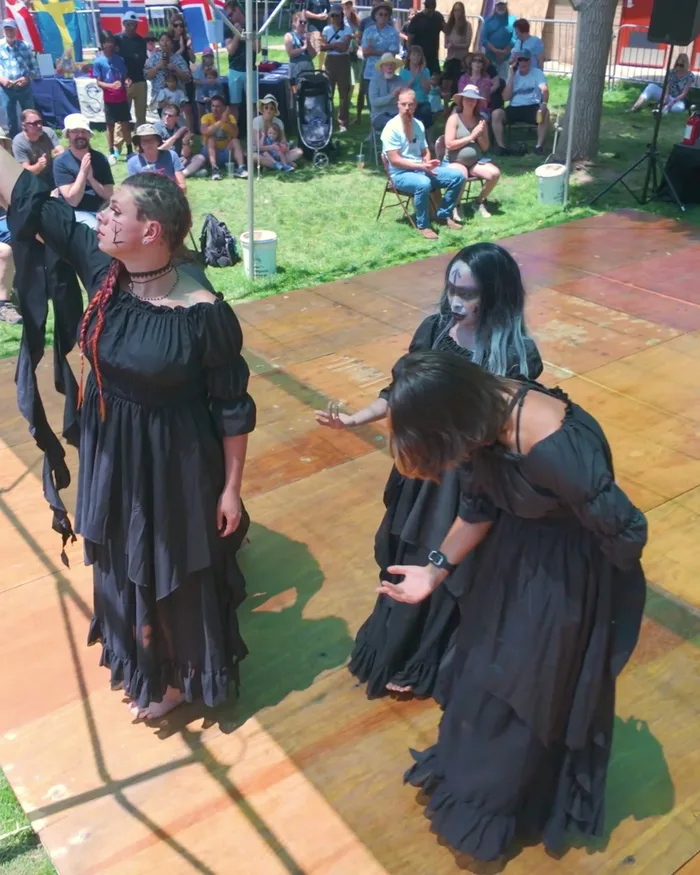 Dancers in black with pale face paint gathered close together on the stage, one reaching a hand skyward