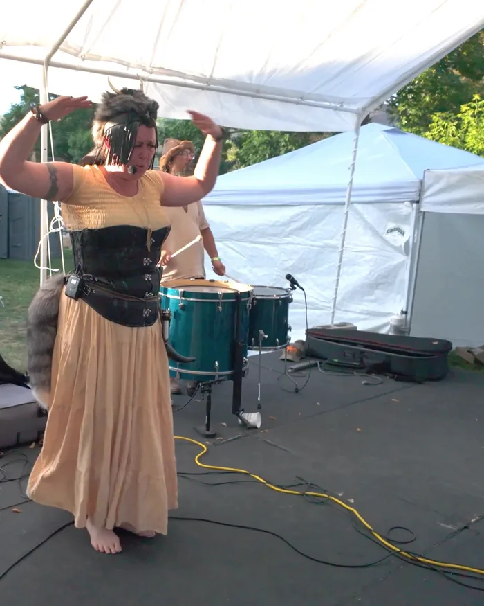 Nordic Daughter performing under the festival tent at the Scandinavian Midsummer Festival — the singer with arms raised, the drummer and guitarist beside her, Nordic flags overhead