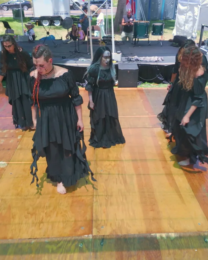 All five dancers in flowing black dresses and pale face paint moving together across the wooden stage, Nordic flags and the crowd behind them