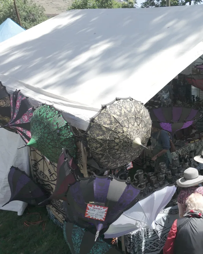 Colorful patterned parasols hanging from a market tent's outer edge in full sun, blue sky and the Golden foothills above, a shopper browsing a table of pewter goblets behind them