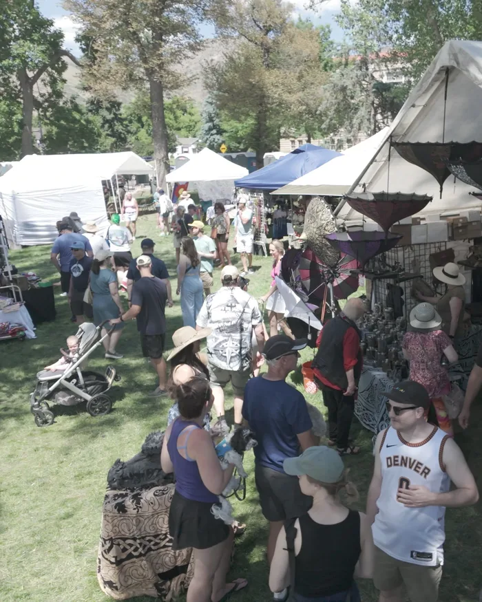 A market row stall selling hats and pewter goblets, a dense crowd of festivalgoers browsing the tables in the sun