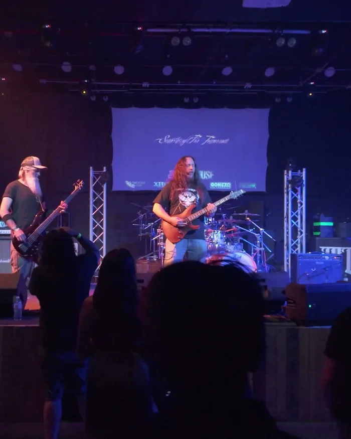 The bassist and lead guitarist of Something for Tomorrow performing side by side at The Rickhouse under blue stage light, a small crowd watching from the floor