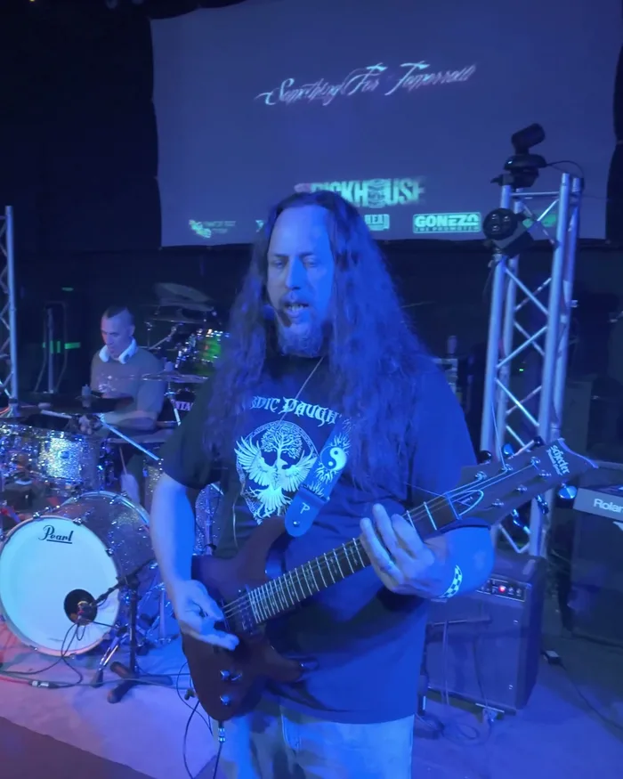Something for Tomorrow's Jason Lycan singing and playing guitar at The Rickhouse, the venue's screen displaying the band's name above the stage, the drummer visible behind him