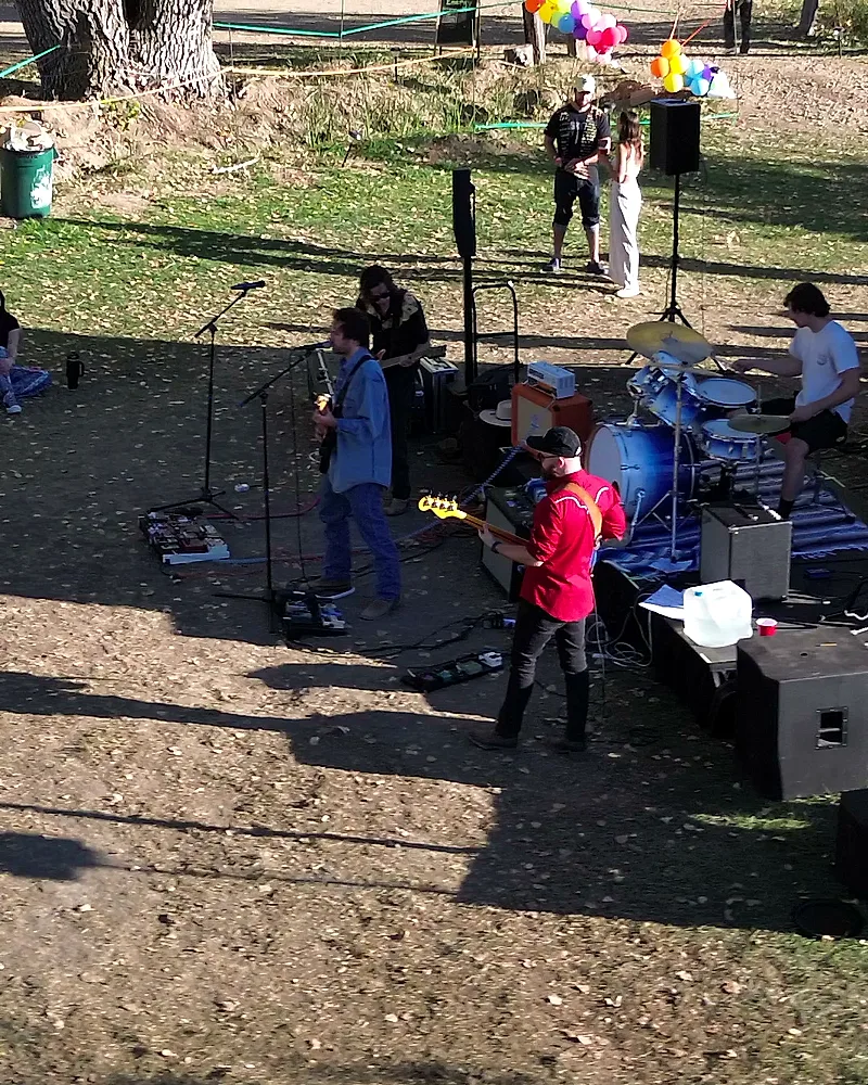 An aerial view over the Sunday stage — the full band performing, two festivalgoers standing together near a rainbow balloon display, parked cars and cottonwood trees in the grounds beyond