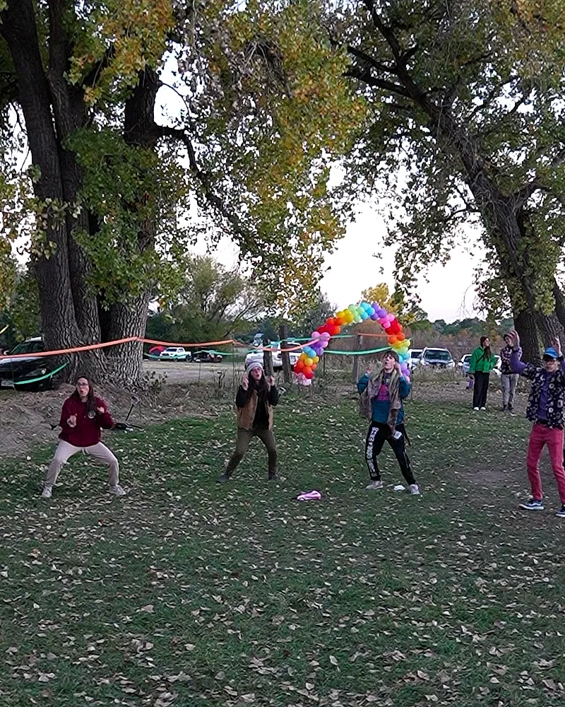 Several festivalgoers dancing together in the grass under a full rainbow balloon arch, cottonwoods and parked cars in the grounds beyond, Sunday afternoon
