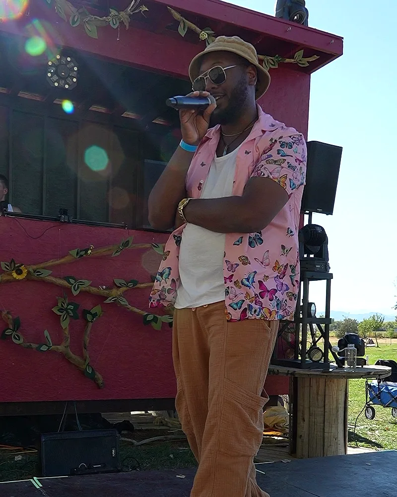 The festival's emcee, sunglasses and a bucket hat, addressing the crowd on the mic beside the red stage trailer, Front Range foothills behind