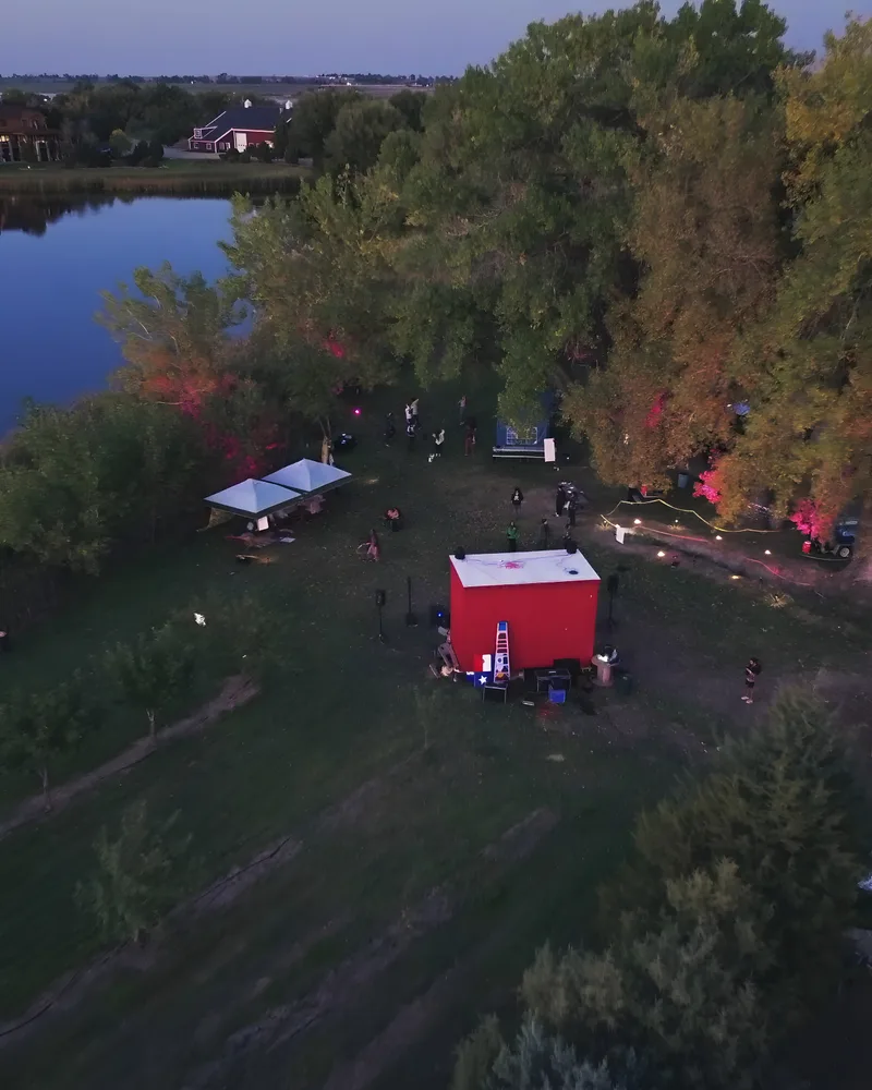 A blue-hour aerial reveal over the Vybe festival grounds beside a lake, string lights glowing through the cottonwoods