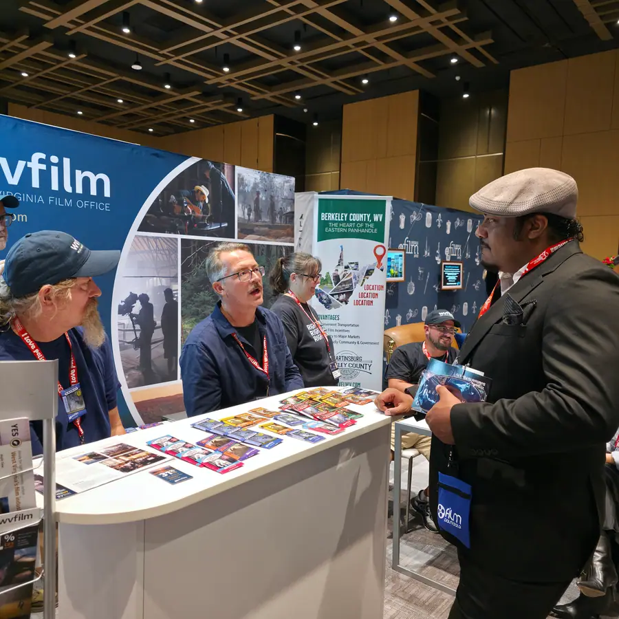 Two attendees talking with representatives at the West Virginia Film Office booth, browsing location photos and brochures at their market table.