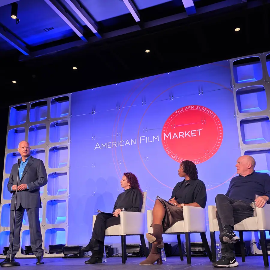 A presenter standing at a microphone on the left of the AFM Sessions stage, three listeners seated in chairs to his right.