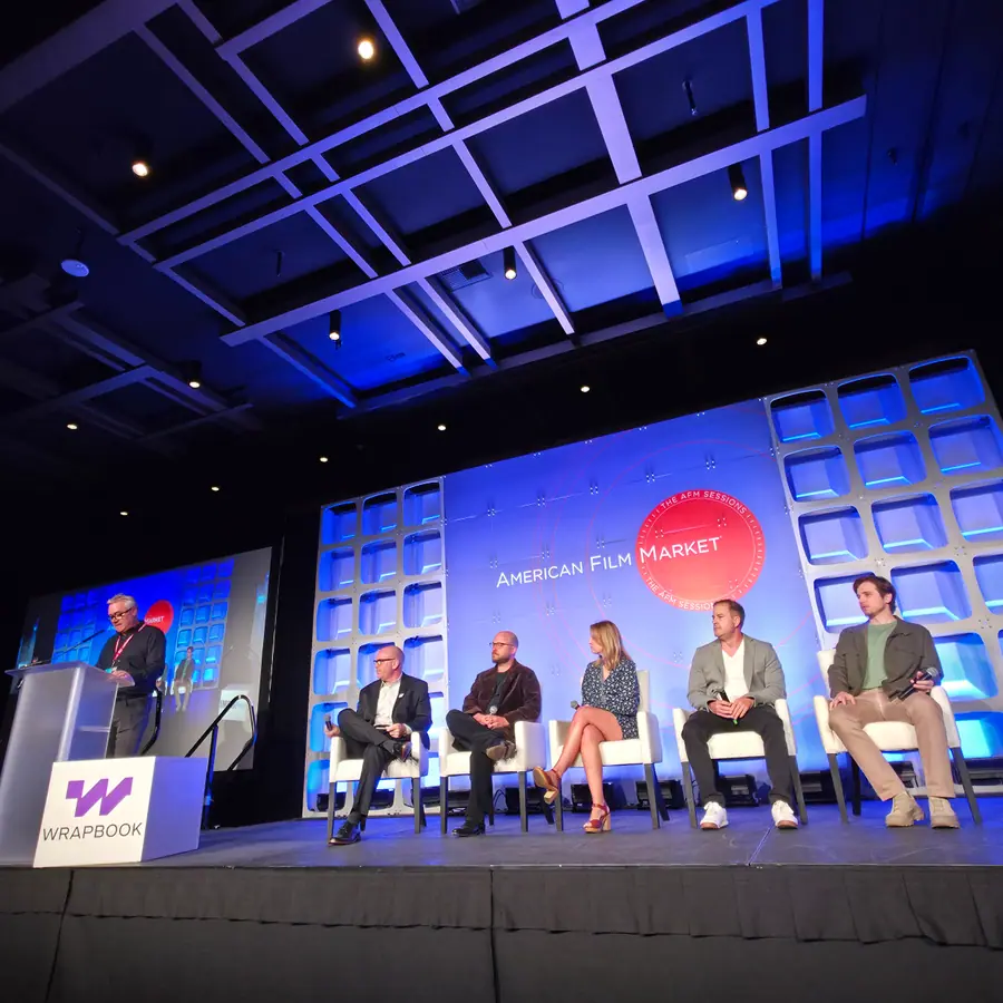 The same stage from further back — a moderator at a sponsor lectern on the left, five panelists seated under the American Film Market backdrop.