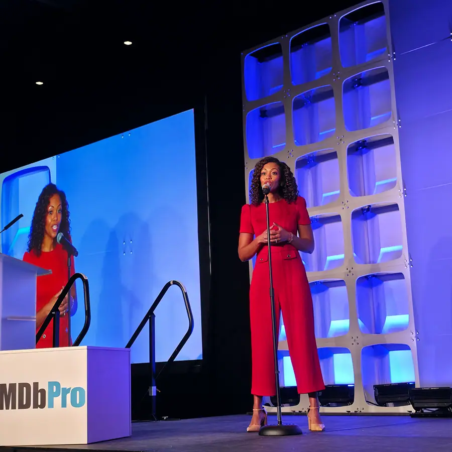A presenter in a red jumpsuit speaking at a standing microphone beside the sponsor podium, her image mirrored on the big screen behind her.