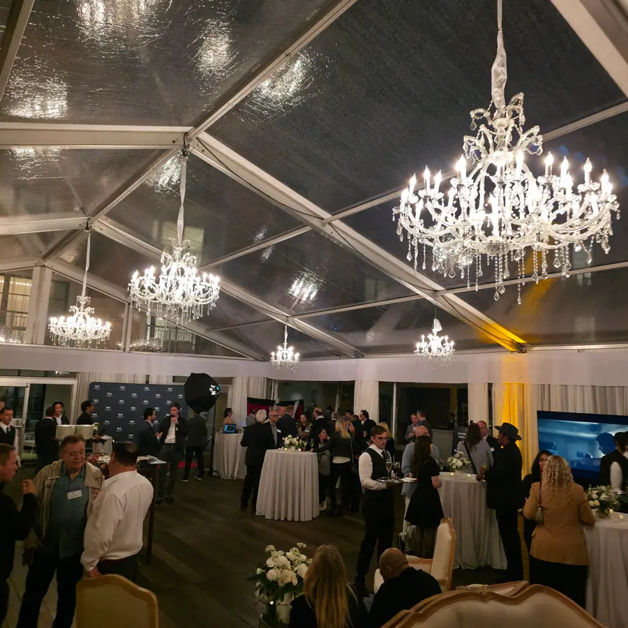 Guests mingling at an evening marquee reception under chandeliers, a step-and-repeat backdrop visible in the background.