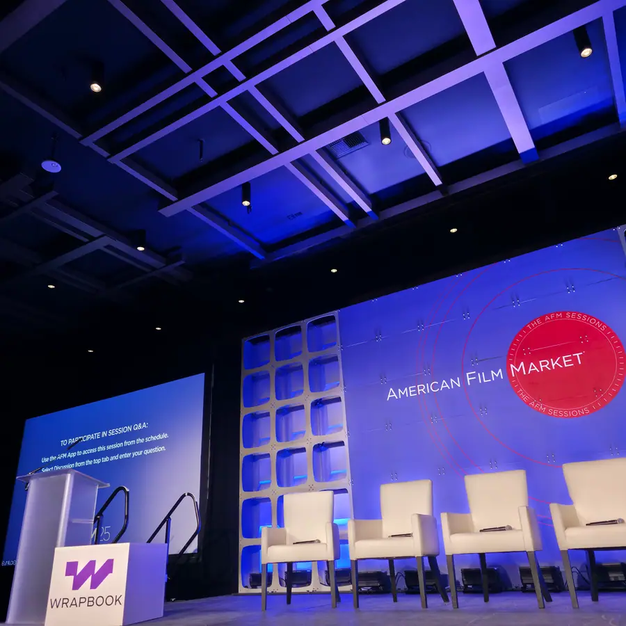 An empty conference stage under blue lighting, American Film Market branding on the backdrop, five white chairs set out and a side screen showing session Q&A instructions.