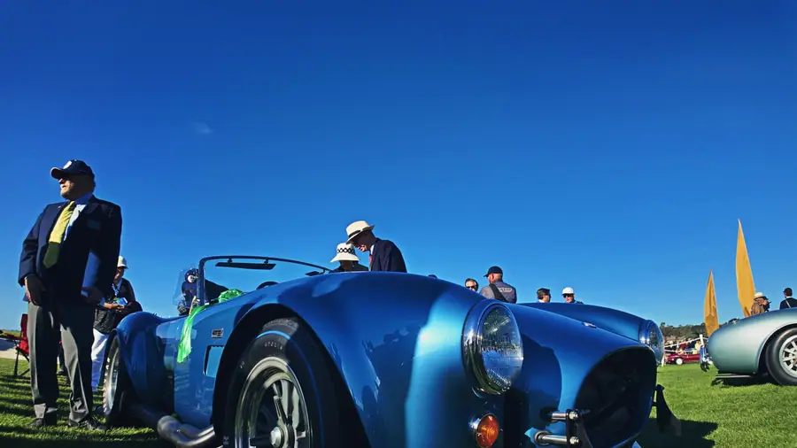 Front three-quarter view of a blue Shelby Cobra with a green ribbon on the fender under a clear sky, a pale Cobra visible in the background