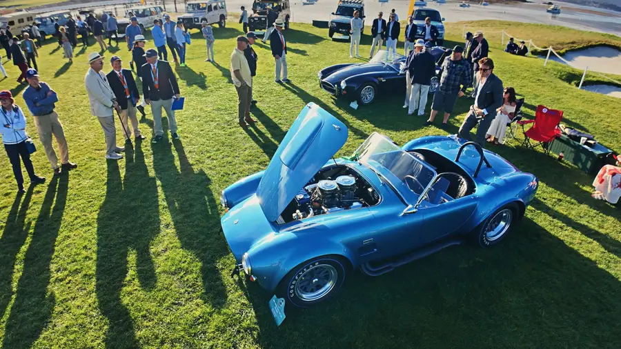 Close view into the open engine bay of a blue Shelby Cobra showing twin carburetors, a crowd of onlookers visible in the background