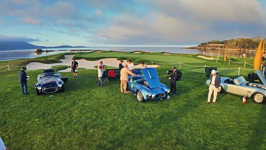 Wide view of Pebble Beach's 18th green at sunrise, three Shelby Cobras spaced across the fairway, bunkers and the bay behind, orange marker flags in the water