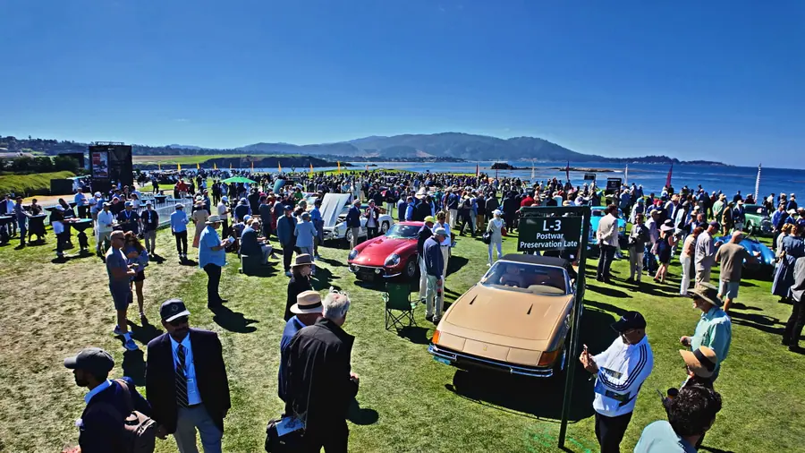 A dense midday crowd on the Pebble Beach show field, a class sign reading "L-3 Postwar Preservation Late," coastline behind