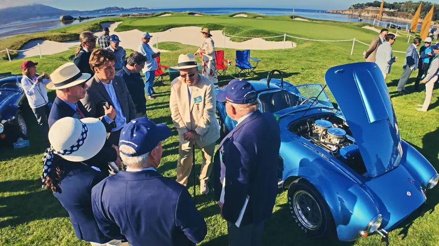 A blue Shelby Cobra with its hood up on a golf fairway at dawn, a small group of people around it, the bay and coastline behind