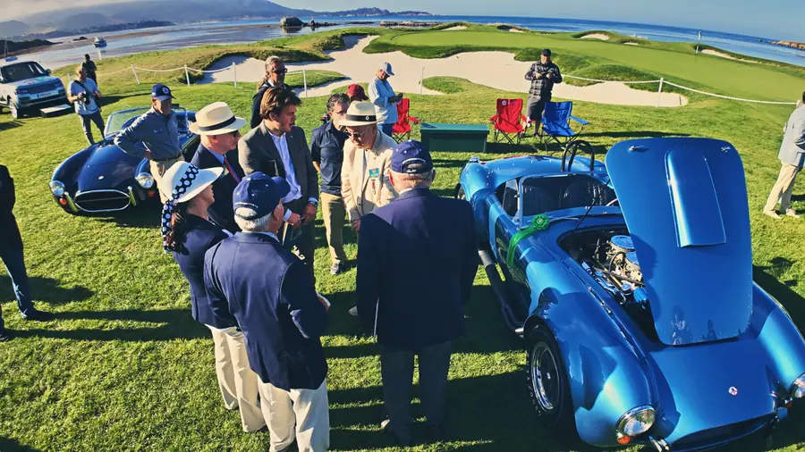 A blue Shelby Cobra with a green class ribbon on its front fender, a small crowd of spectators gathered around it on the show lawn