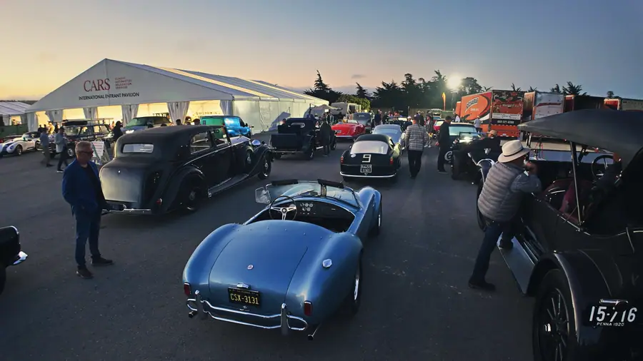The interior of a large event tent marked CARS — Classic Automotive Relocation Services, International Entrant Pavilion, postwar sedans and a pale blue Cobra parked inside at dawn