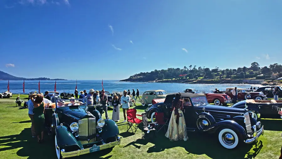 A line of black pre-war luxury cars parked along the Pebble Beach coastline, well-dressed spectators including a woman in a gold gown, the Pacific behind