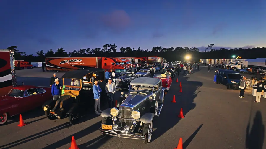A row of car-haulers and already-parked pre-war classics in a floodlit staging lot before sunrise, orange cones marking the lanes