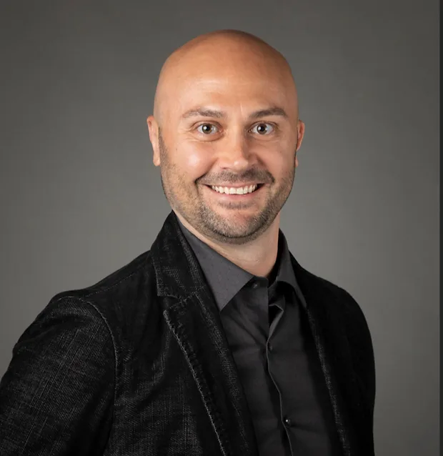 Studio portrait of Adam Smestad, bald, short beard, smiling, black blazer, gray backdrop.
