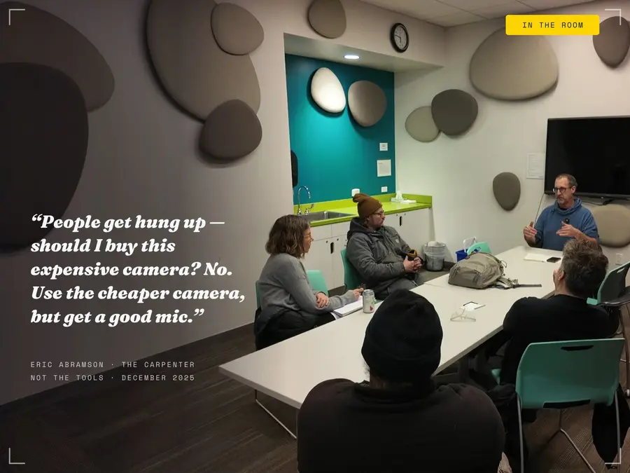 Eric Abramson teaching The Carpenter Not the Tools, gesturing at the head of a long table as attendees listen; white text composited over the room's left wall carries his quote: "People get hung up — should I buy this expensive camera? No. Use the cheaper camera, but get a good mic."