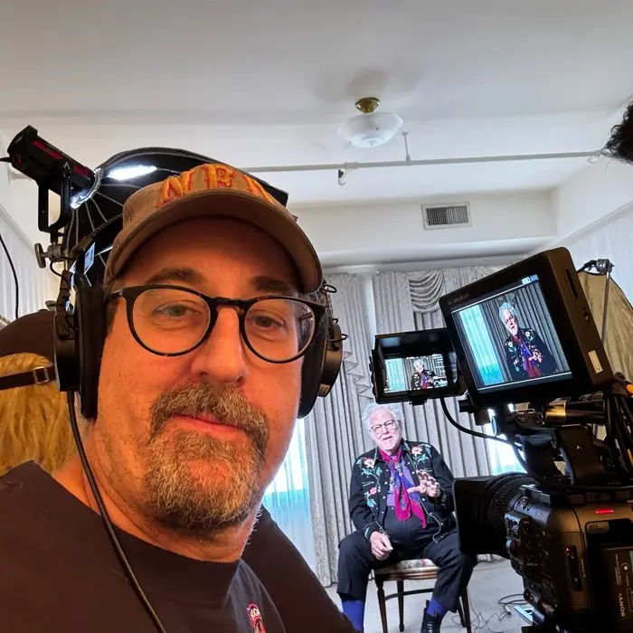 Eric Abramson on a shoot — ballcap, glasses and headphones, a camera rig and two monitors beside him, and the interview subject lit and seated behind.