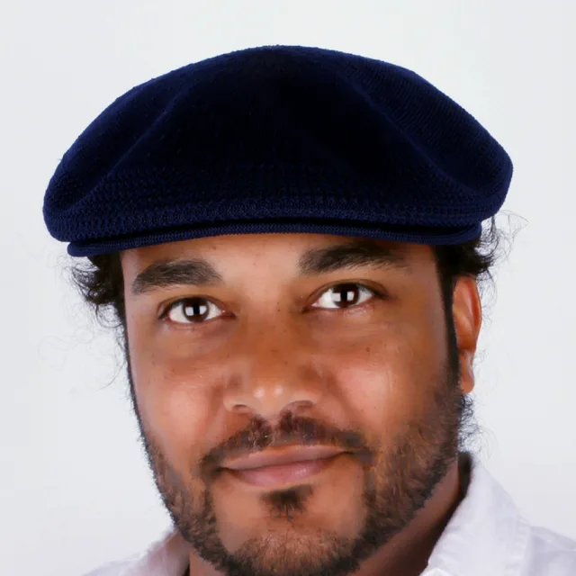 Close portrait of Nick C. Goins Jr wearing a navy flat cap, white shirt, looking directly at camera.
