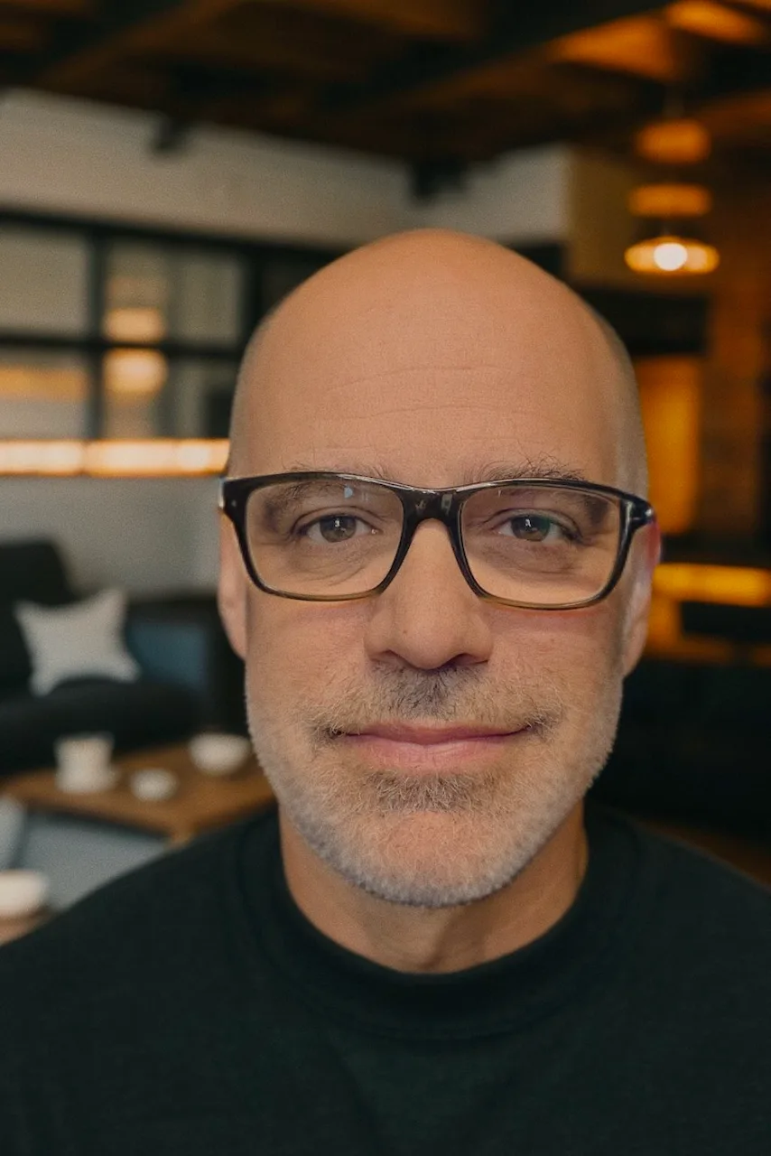 Portrait of Steve Borne, bald, glasses, gray beard, dark sweater, softly lit interior background.