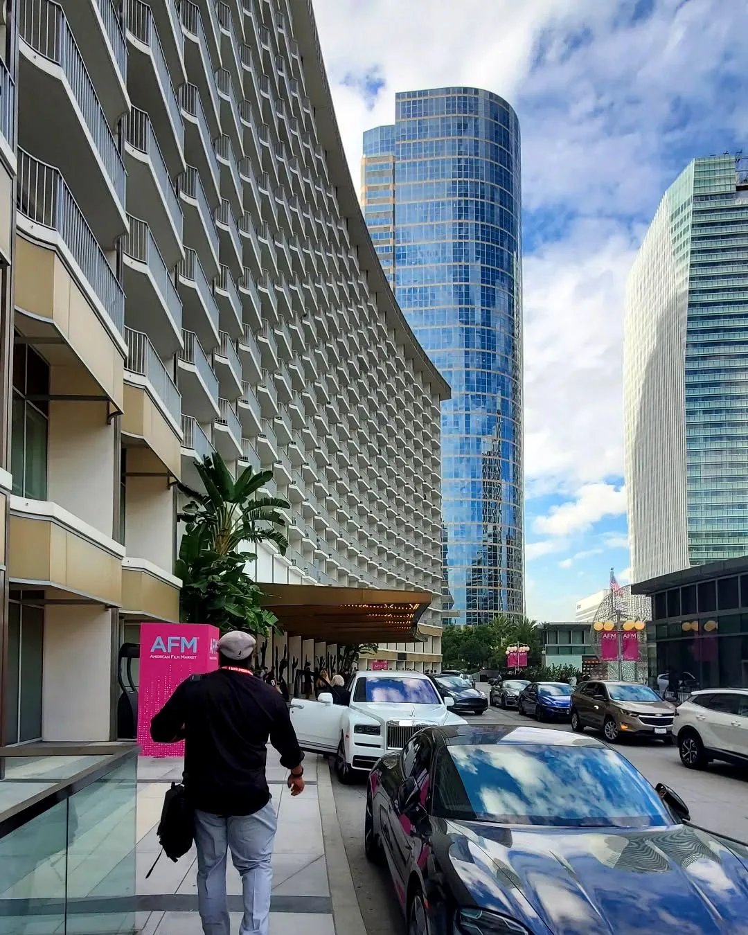 The approach to the American Film Market in Los Angeles: a curved hotel facade sweeping down to its entrance, glass towers and cloud behind it, pink AFM banners along the arrival lane, one attendee walking in away from camera.