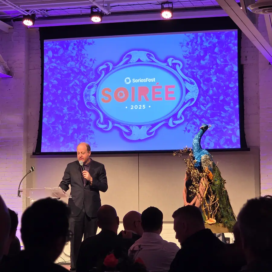 Governor Jared Polis addressing the room at the SeriesFest Soirée, a ceramic peacock installation beside the stage.