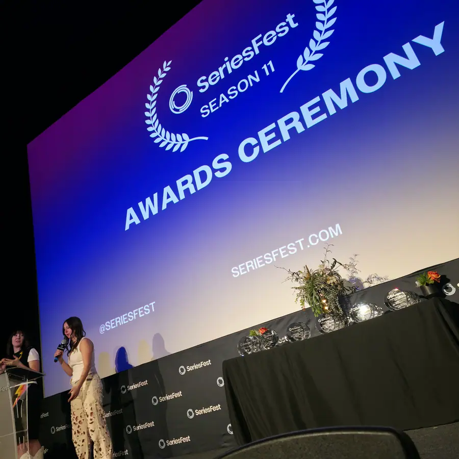 A host at the podium during the Season 11 Awards Ceremony, the ceremony titlecard filling the screen behind the stage.