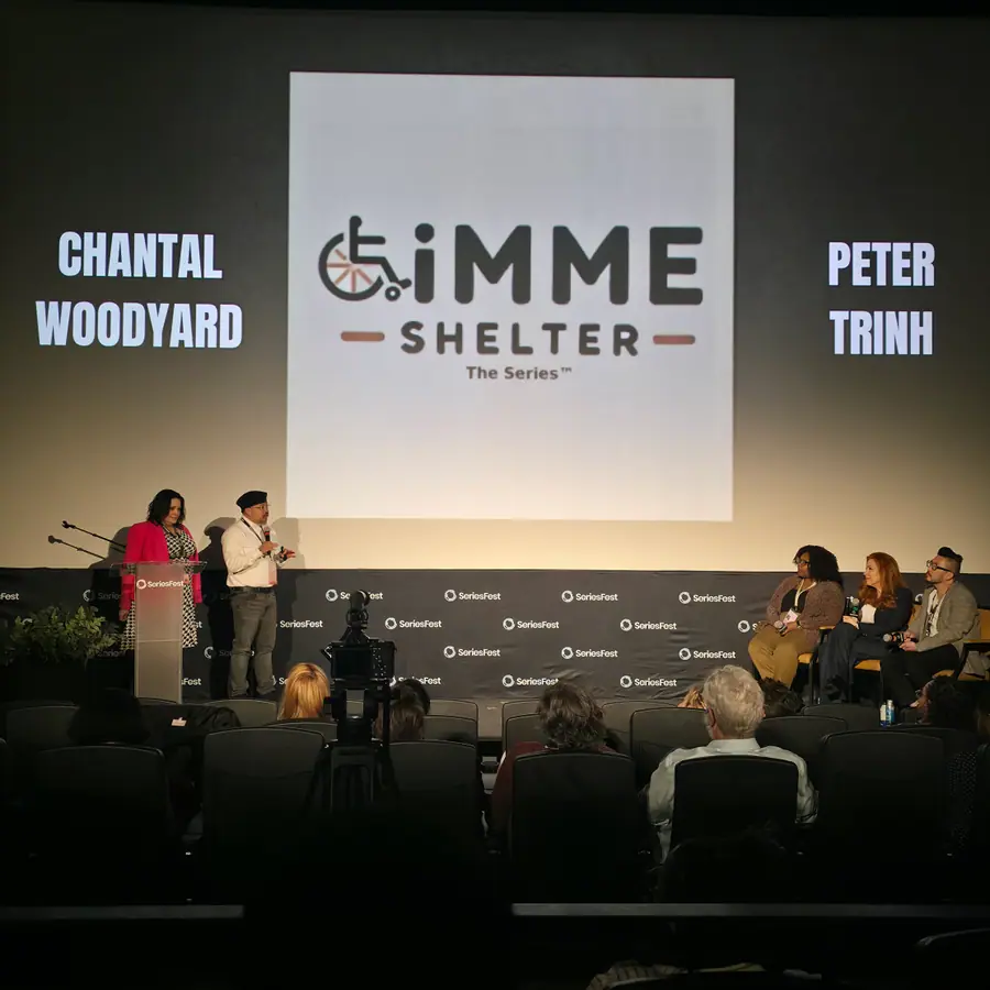 Two people at the podium beside a panel row, the screen behind them showing the Gimme Shelter (The Series) logo with the names Chantal Woodyard and Peter Trinh.