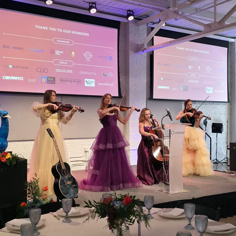 A string quartet in formal gowns performing on the Soirée stage, the sponsor wall projected behind them.