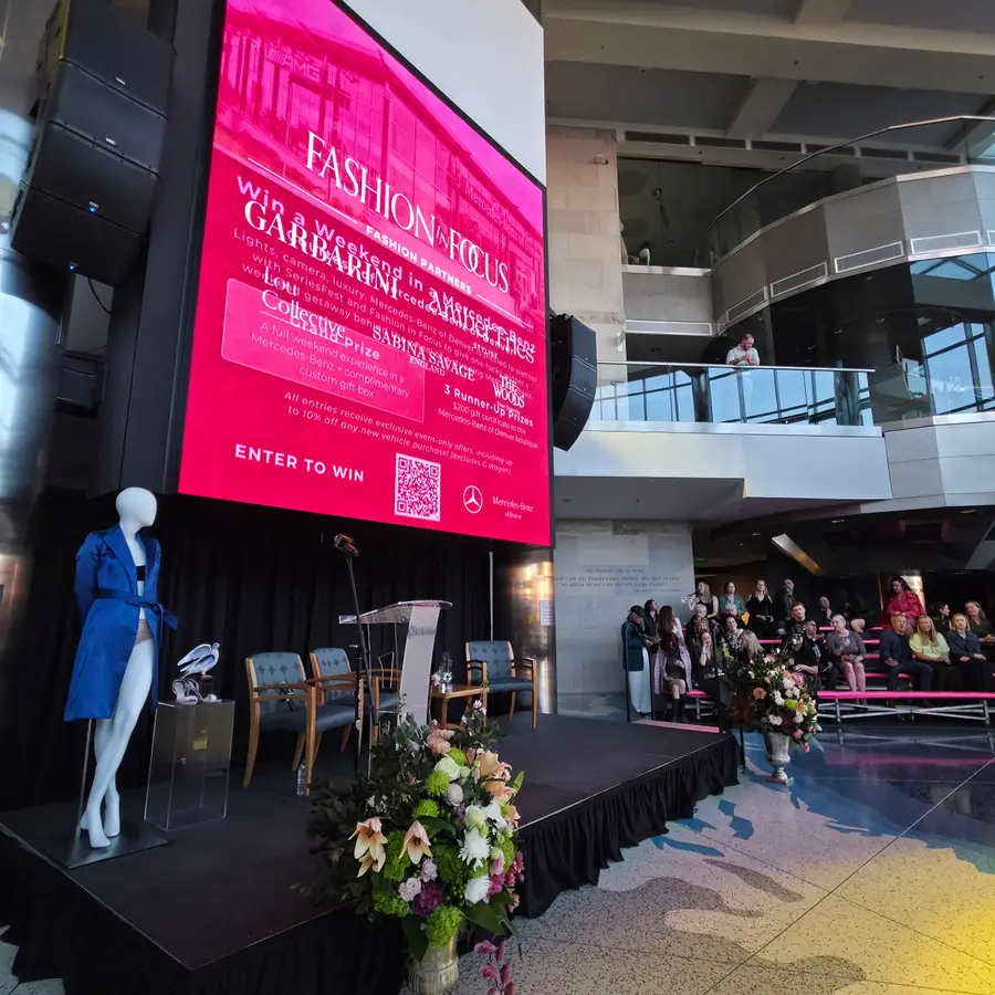 The Fashion in Focus title screen at The Cable Center, a blue coat on a mannequin staged beside the podium, the runway audience seated behind.